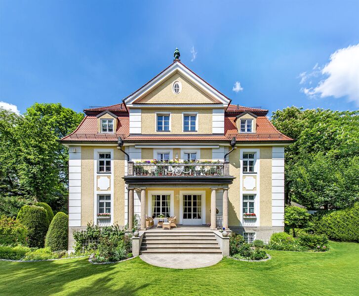 Yellow rendered villa with a mansard roof, balconies and a curved exterior staircase, surrounded by lawn and topiary.