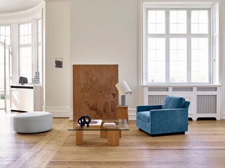 Blue upholstered armchair with an angular backrest next to a low wooden table in front of a bright wall of windows.