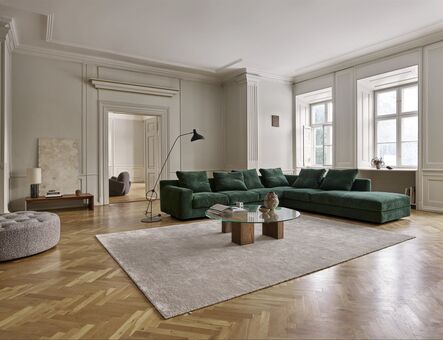 Dark green corner sofa on a light rug in a period room with herringbone parquet and stucco mouldings.