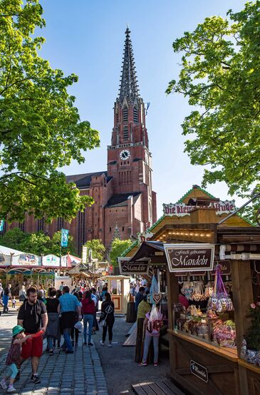 Marktbuden und ein Fahrgeschäft auf einem Platz, dahinter der hohe Backsteinturm der Mariahilfkirche.