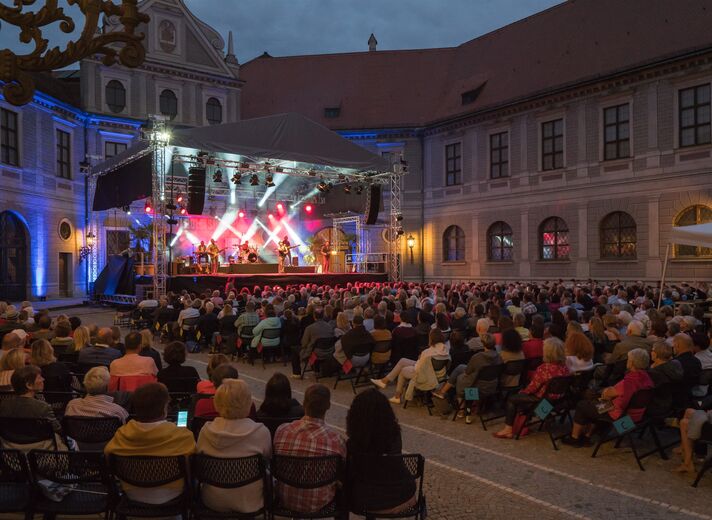 Bühne mit farbigem Scheinwerferlicht vor historischer Fassade, davor Publikum auf Stuhlreihen in der Dämmerung.