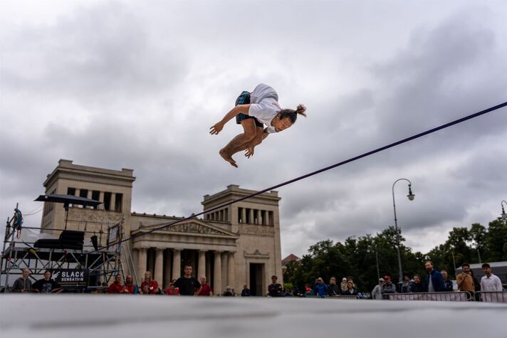 Ein Teilnehmer balanciert auf einer Slackline über einer Wiese, ringsum Zuschauer und Sportstände.