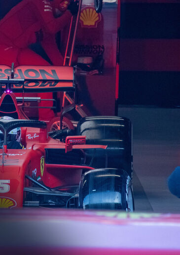 Red Formula 1 car in the pit garage, mechanics and advertising boards in the background.