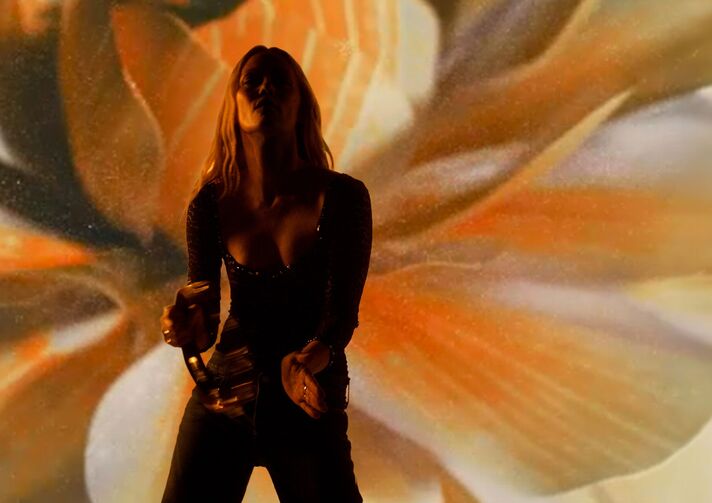 Singer with a microphone backlit on a stage, behind her a large projection of blossoms in shades of orange.