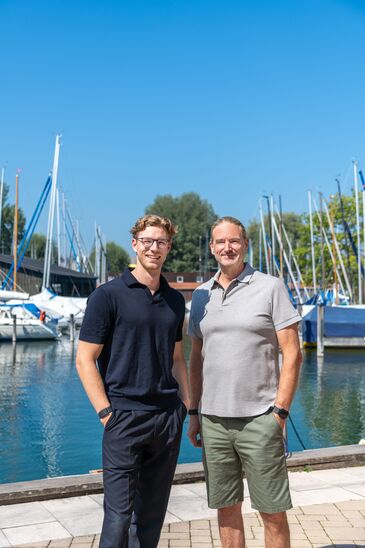 Zwei Männer stehen nebeneinander am Steg, hinter ihnen liegen Segelboote mit aufgerichteten Masten.