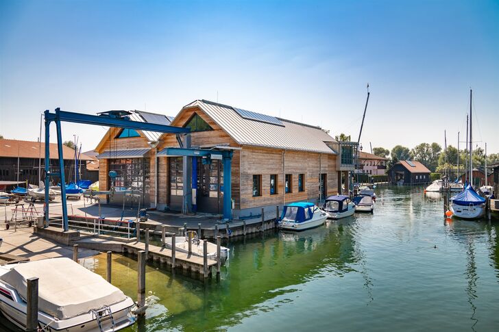Werftgebäude aus Holz mit blauem Portalkran am Wasser, davor liegen Motorboote im Hafenbecken.