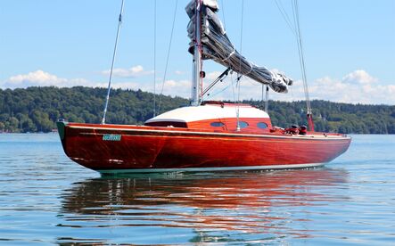 Rot lackierte Segelyacht mit geborgenem Segel liegt auf dem ruhigen See, dahinter ein bewaldetes Ufer.