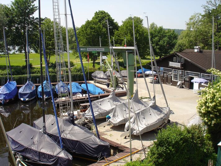 Auf dem Werftgelände stehen abgedeckte Jollen und Yachten an Land, dahinter das dunkle Werftgebäude am Seeufer.