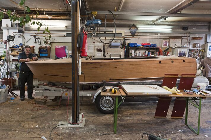 Ein Bootsbauer arbeitet in der Halle an einem aufgebockten Holzmotorboot, dessen Rumpf blank geschliffen ist.