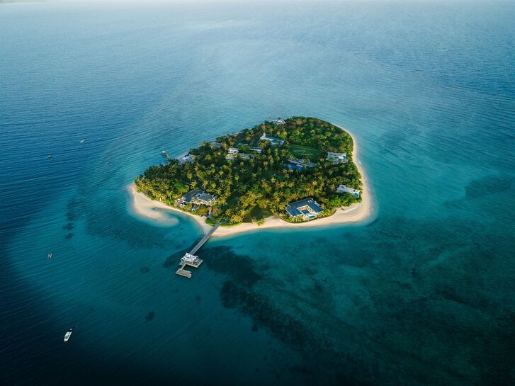 Small green island with a light sandy beach and a boat jetty, surrounded by turquoise shallows and deep blue sea.