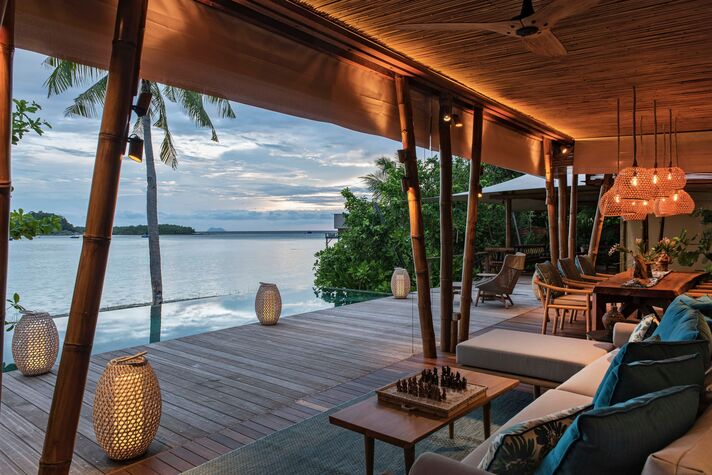 Open lounge under a high wooden roof with armchairs, lanterns and a view across a lagoon to wooded islands.