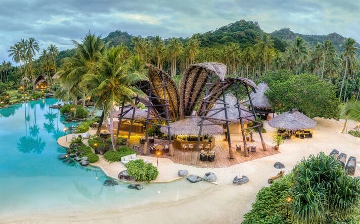 Expansive pool landscape with a sandy beach and a bar under a curved palm-leaf roof, with palm trees and wooded hills behind.