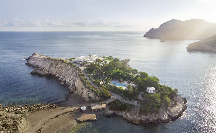 View across a bay to a vegetated rocky island with a villa complex and pool, with open sea and further islands behind in the evening light.