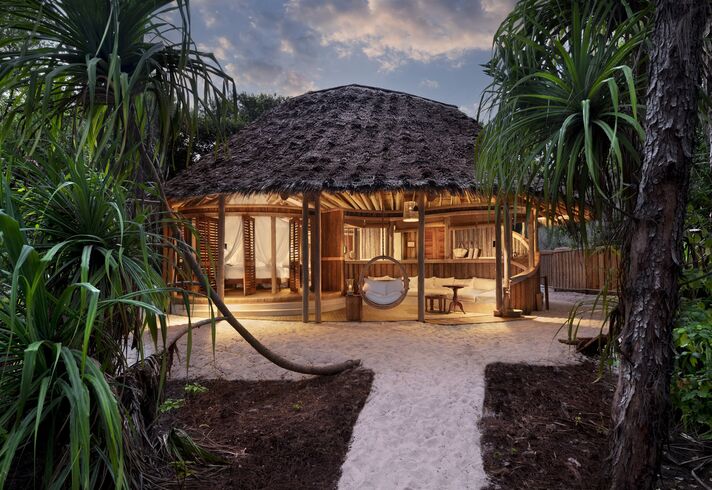 Open bungalow with a round thatched roof amid tropical plants, with a sandy path in front and soft evening lighting.