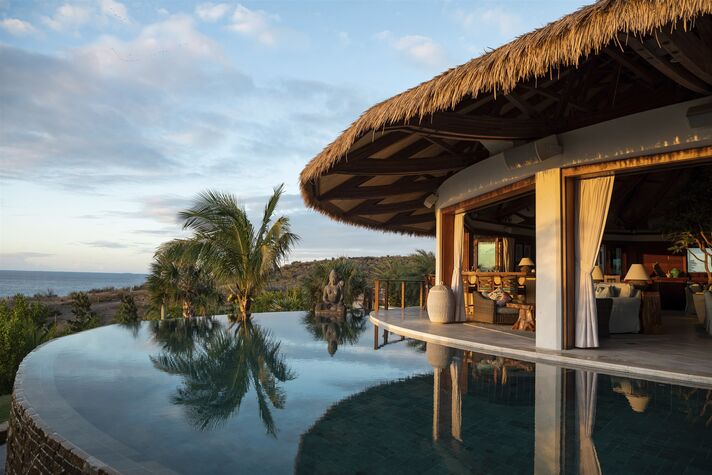 Infinity pool in front of an open building with a palm-leaf roof, with palm trees and the sea at sunset in the background.