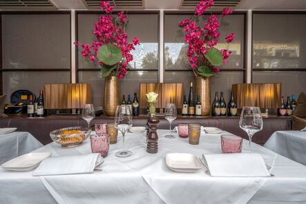 Festlich eingedeckter Tisch mit rosa Gläsern und Blumenschmuck vor einer Wand mit Weinregalen.