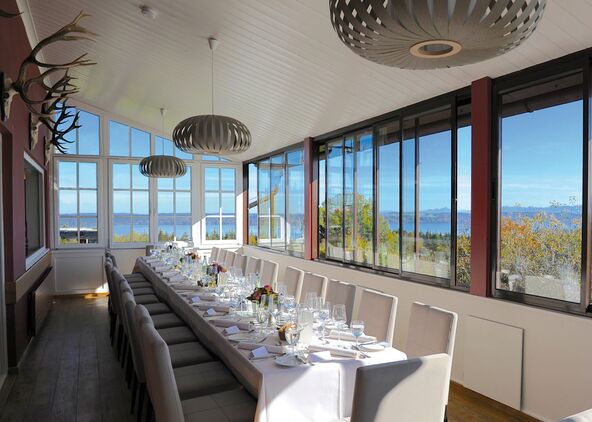 Lange festlich gedeckte Tafel in einem hellen Saal, dessen Fensterfront den Blick über den See freigibt.