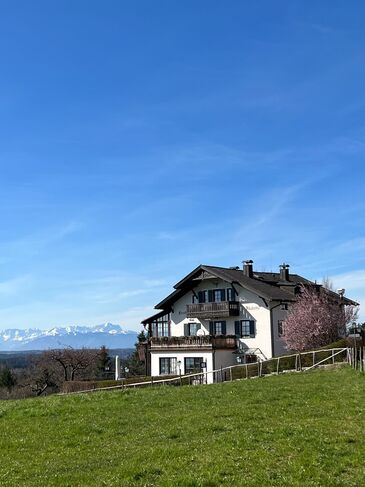 Weiß verputztes Landhaus mit Krüppelwalmdach steht auf einer Wiesenkuppe, dahinter zeichnet sich die Alpenkette ab.