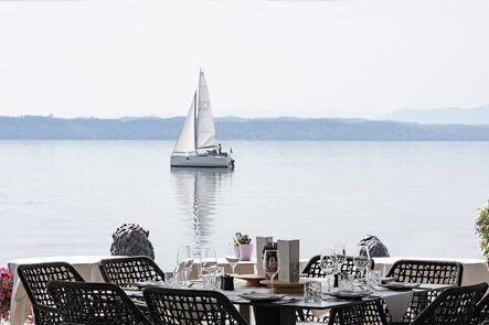 Gedeckte Tische mit dunklen Korbstühlen auf einer Terrasse am Wasser, im Hintergrund liegt ein Segelboot auf dem See.