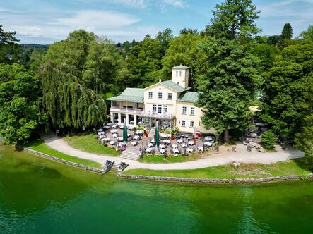 Luftaufnahme eines weißen Gasthauses mit Türmchen direkt am bewaldeten Ufer, davor Sonnenschirme und ein Steg im türkisen Wasser.