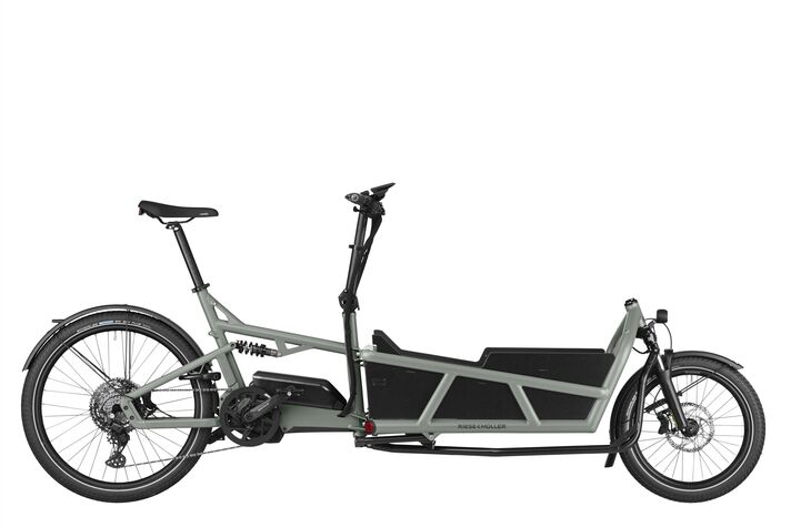 Dark grey electric cargo bike with a low transport box in front of the front axle and a display on the handlebars, side view against a pale wall.