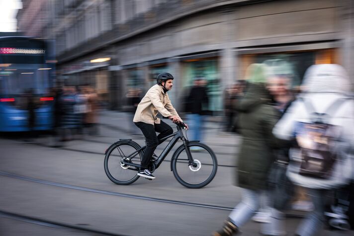 Cyclist on a slim e-bike with no visible motor on a road, a blurred row of houses in the background.