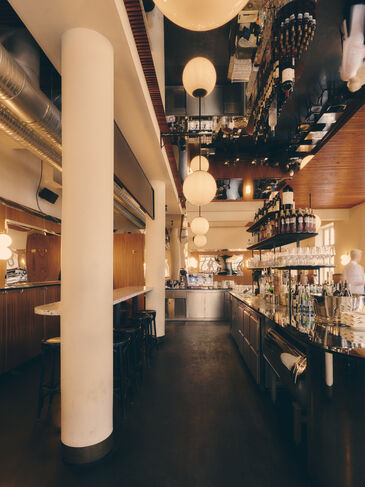 Long bar area with a light round column, globe lights and a bottle shelf, with tables and dark chairs in front in warm light.