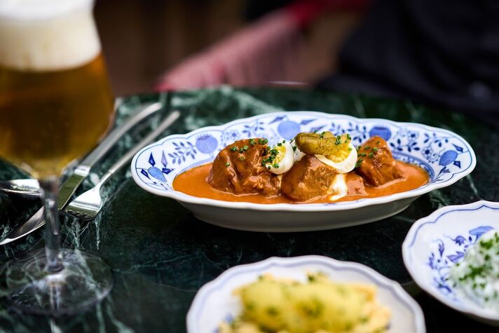 Plate of goulash, Nockerl dumplings and sour cream on blue patterned porcelain, next to a glass of beer on marble.
