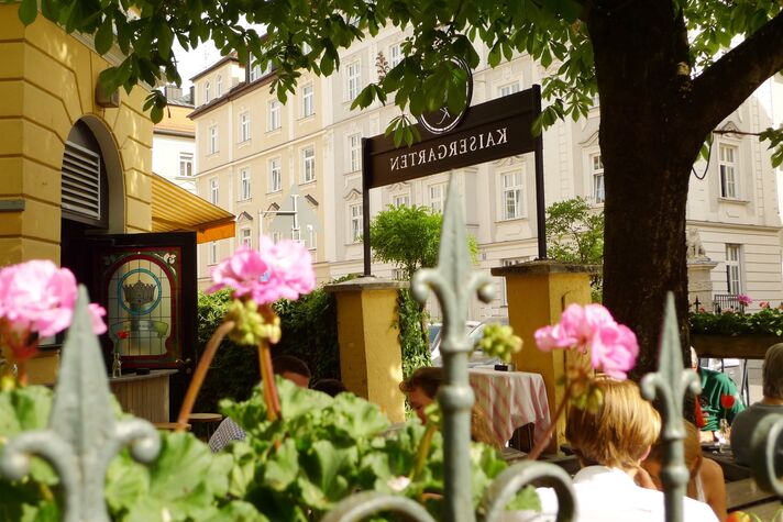 View over flowering geraniums into a shady beer garden with guests at laid tables under old chestnut trees.