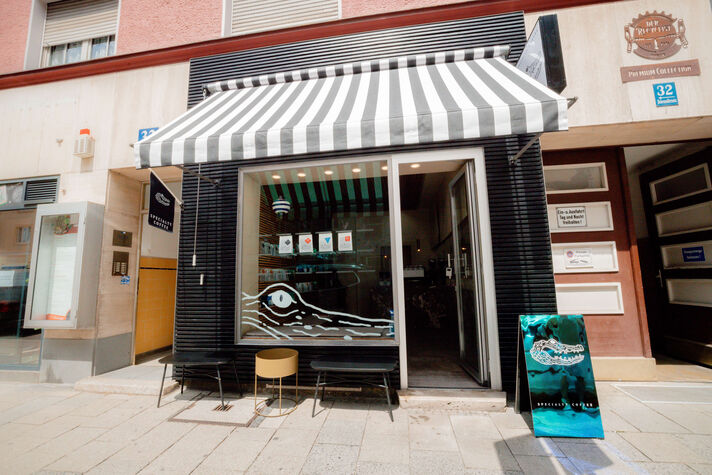 Shopfront of a coffee roastery with a black-and-white striped awning, an open door and bar stools along the window.