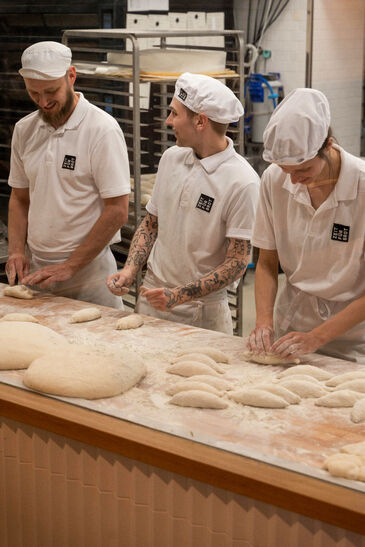 Three bakers in white shirts and headwear shaping dough pieces at a work table, with trays of bread in the background.