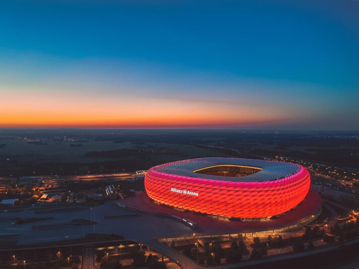 Rot leuchtende Stadionhülle bei Nacht, umgeben von den Lichtern der Zufahrtsstraßen.