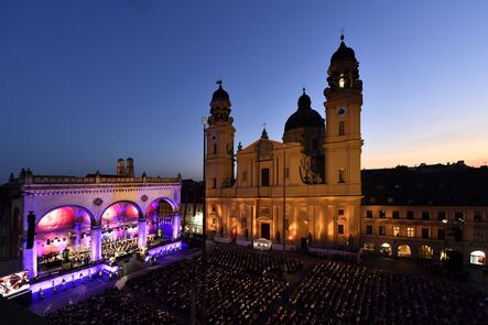 Blau angestrahlte Bühne vor der Theatinerkirche, davor dicht besetzte Stuhlreihen in der Abenddämmerung.