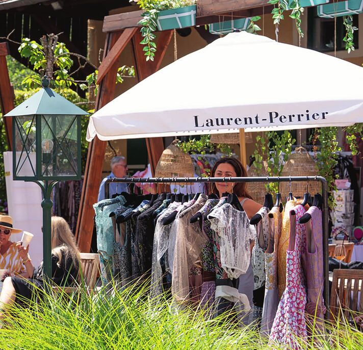 Clothes rail with summer dresses under a white parasol in the garden, behind it visitors among wicker hats and tall grasses.