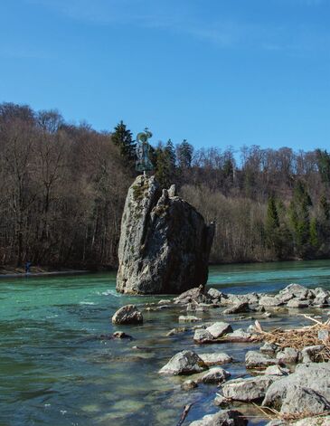 Tall rock with a summit cross rises from the turquoise-green Isar, surrounded by scree in the riverbed and a wooded slope.