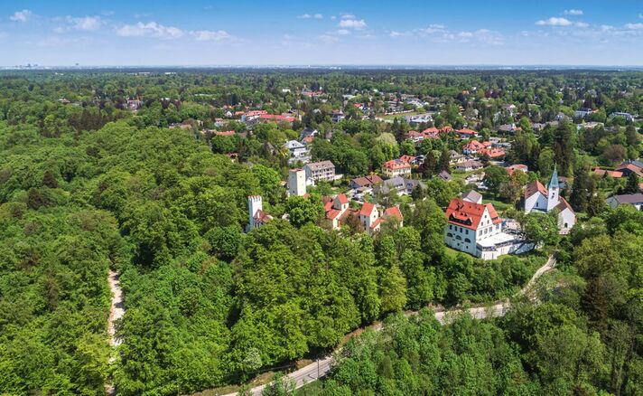 Aerial view over Grünwald with a white church and tower among villa rooftops, the wooded high bank of the Isar in the foreground.