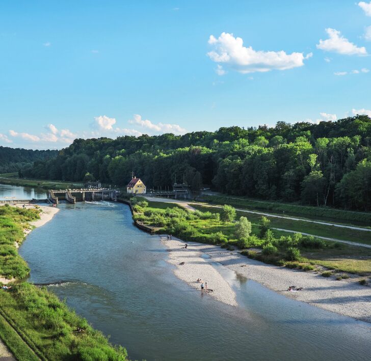 View from above of the Isar with a weir and a yellow keeper’s house, pale gravel banks in the shallow water in front and wooded slopes.