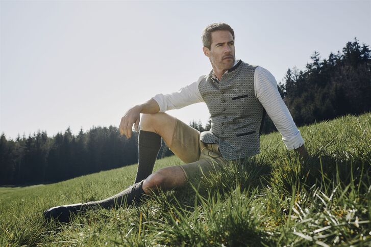Man in light lederhosen, a white shirt and a grey-green gilet kneels in a meadow in front of the edge of a forest.