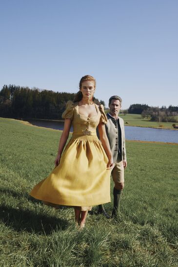 Woman in a bright yellow dirndl walks across a meadow, with a lake and a man in traditional dress in the background.