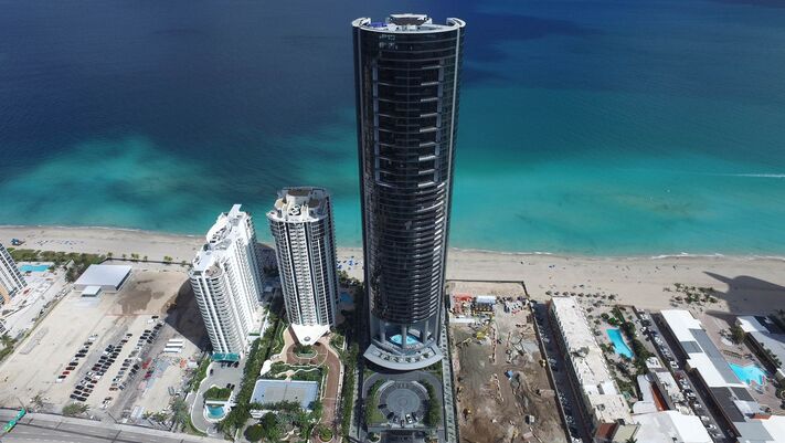 Dunkler Rundturm überragt die flachere Bebauung an einem breiten Sandstrand mit türkisem Wasser.