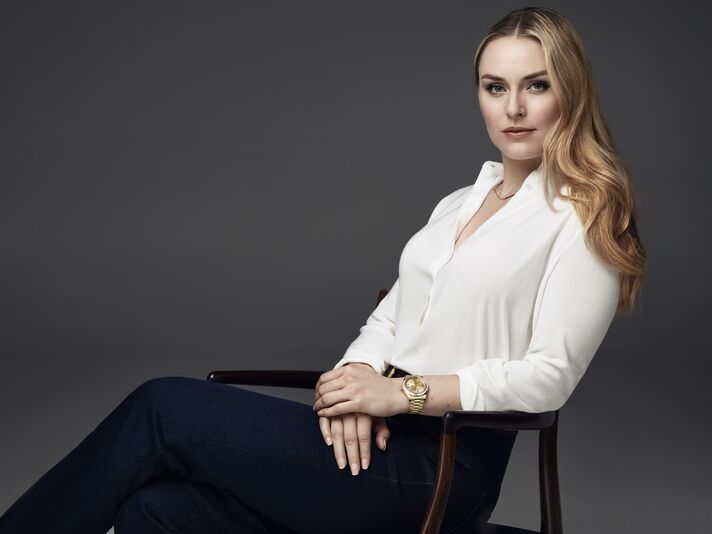 Lindsey Vonn sitzt in weißer Bluse und dunkler Hose auf einem Stuhl vor grauem Hintergrund, am Handgelenk eine goldene Armbanduhr.