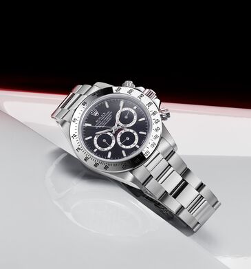 Chronograph aus Edelstahl mit schwarzem Zifferblatt, drei Hilfszifferblättern und Tachymeterskala, liegend auf spiegelnder Fläche.