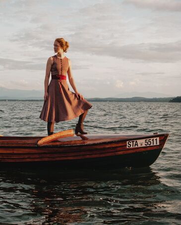 Zwei Dirndl in Burgund und gedecktem Blau hängen an einer Holzstange, daneben Blusen und Schürzen aus gemustertem Stoff.
