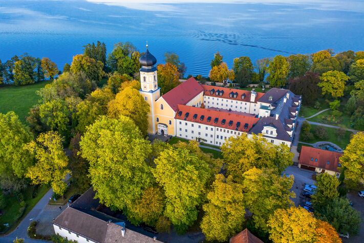 Luftbild der Klosteranlage mit Kirchturm und roten Ziegeldächern, umgeben von altem Baumbestand am Seeufer.