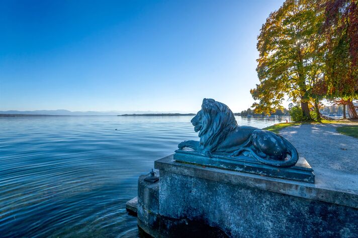 Liegender Löwe aus Stein auf einem Sockel am Seeufer, dahinter das weite Wasser und der herbstliche Baumbestand.