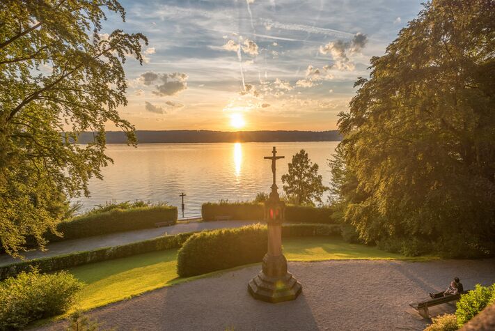 Steinkreuz und Denkmal auf einer gepflegten Uferwiese im Abendlicht, dahinter der See und die untergehende Sonne.