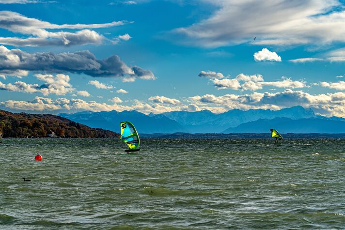Windsurfer mit buntem Segel gleitet über das kabbelige Wasser, am Horizont die Uferlinie und die Alpenkette.