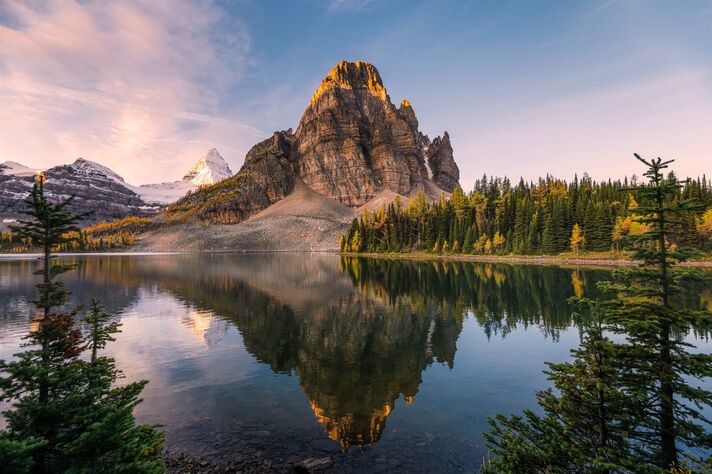 A pointed rocky peak is reflected in a still mountain lake in the morning light, with a few conifers standing on the shore.