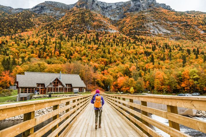 A woman hiker crosses a wide wooden bridge towards a hillside in autumn colours, with a rocky mountain range behind.