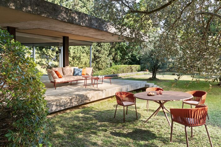 Terrace under a flat canopy with a round dining table, red chairs and lounge furniture, lawn and old trees in front.